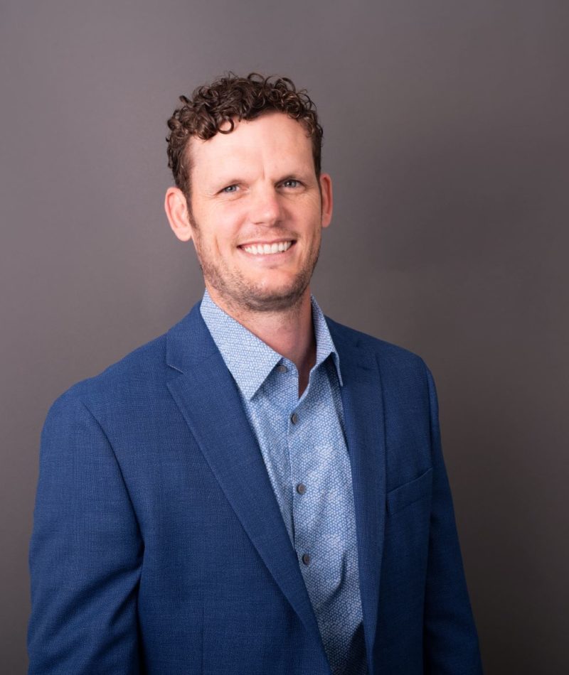 Patrick Hoelscher smiles at the camera. He has a dark blue suite jacket over a casual, light blue button up. He has curly dark hair and hazel eyes.