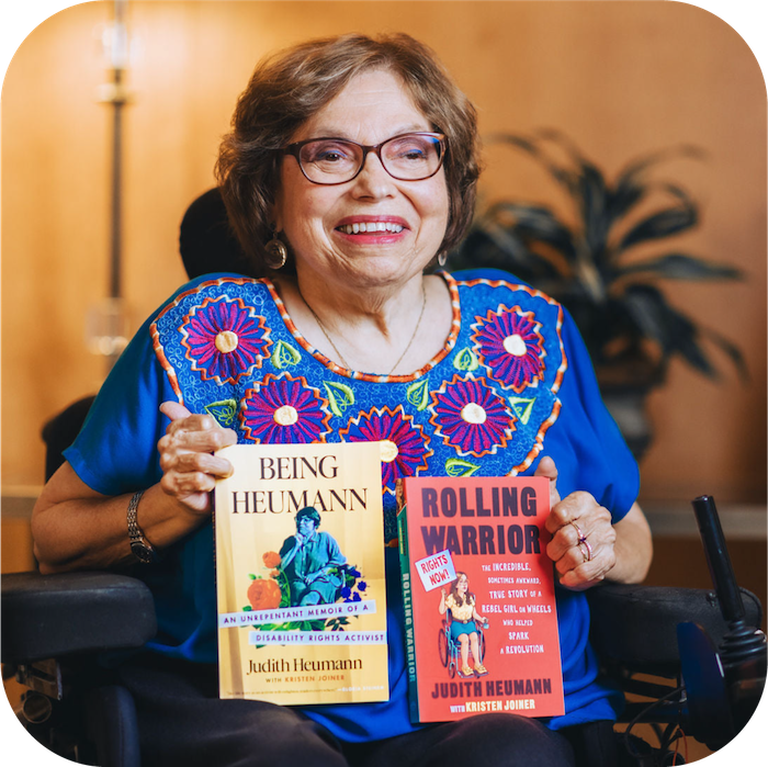 Judy sits in her wheelchair, smiling as she holds two of her books. She wears a blue shirt with purple flowers.