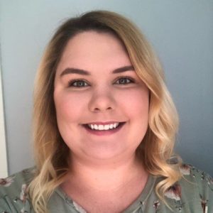 Paige has short curly blond hair and a bright smile. She wears a green floral shirt.