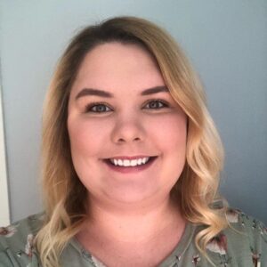 Paige has short curly blond hair and a bright smile. She wears a green floral shirt.