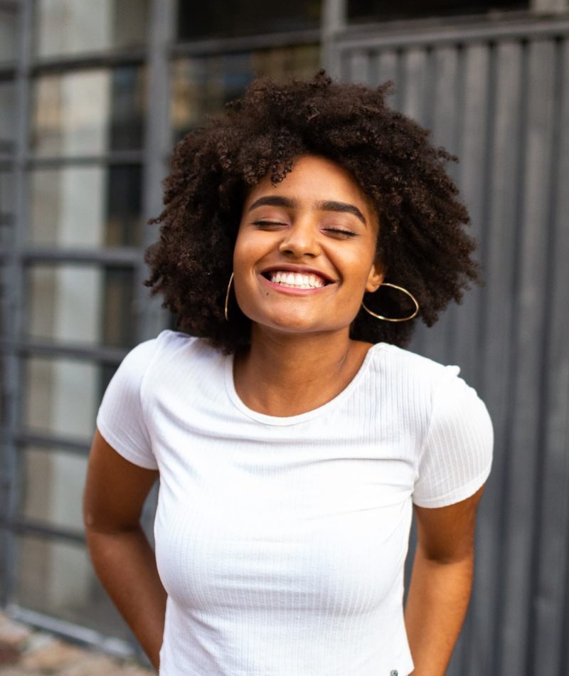 A girl with curly black hair smiles widely, her eyes closed. She has a white shirt on.