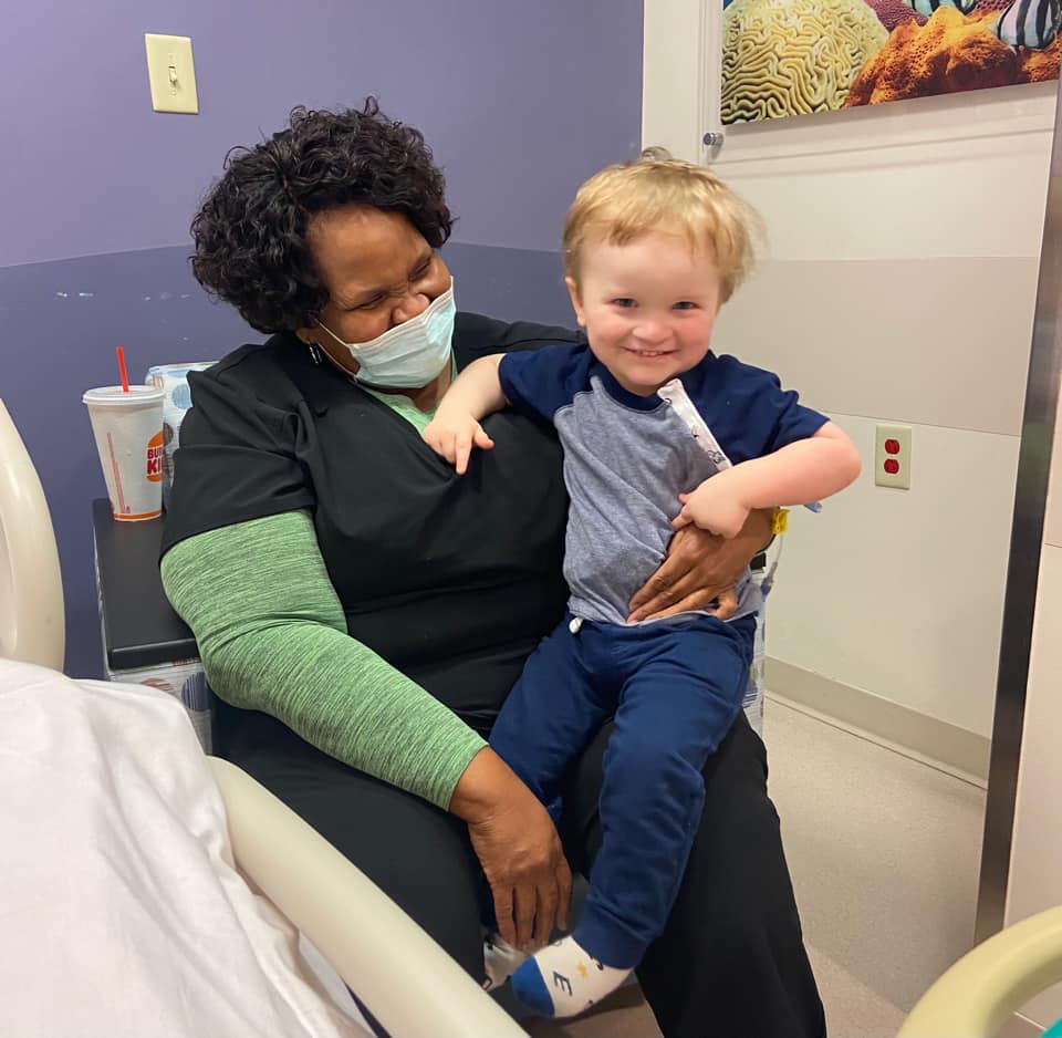 A Black woman with a mask holds a young boy in a hospital room. Both are smiling