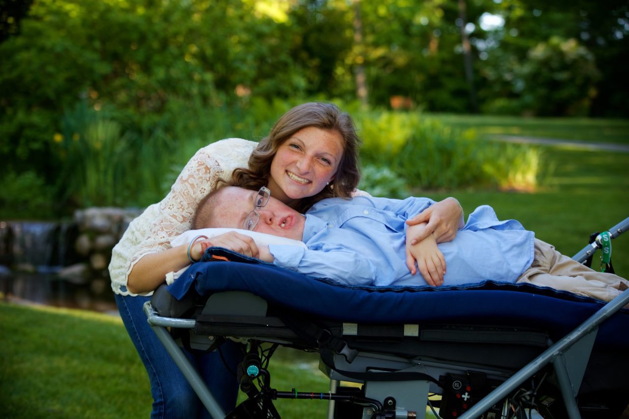 A young woman smiles as she leans in for a picture with a young man with a disability, who lays on a cot.