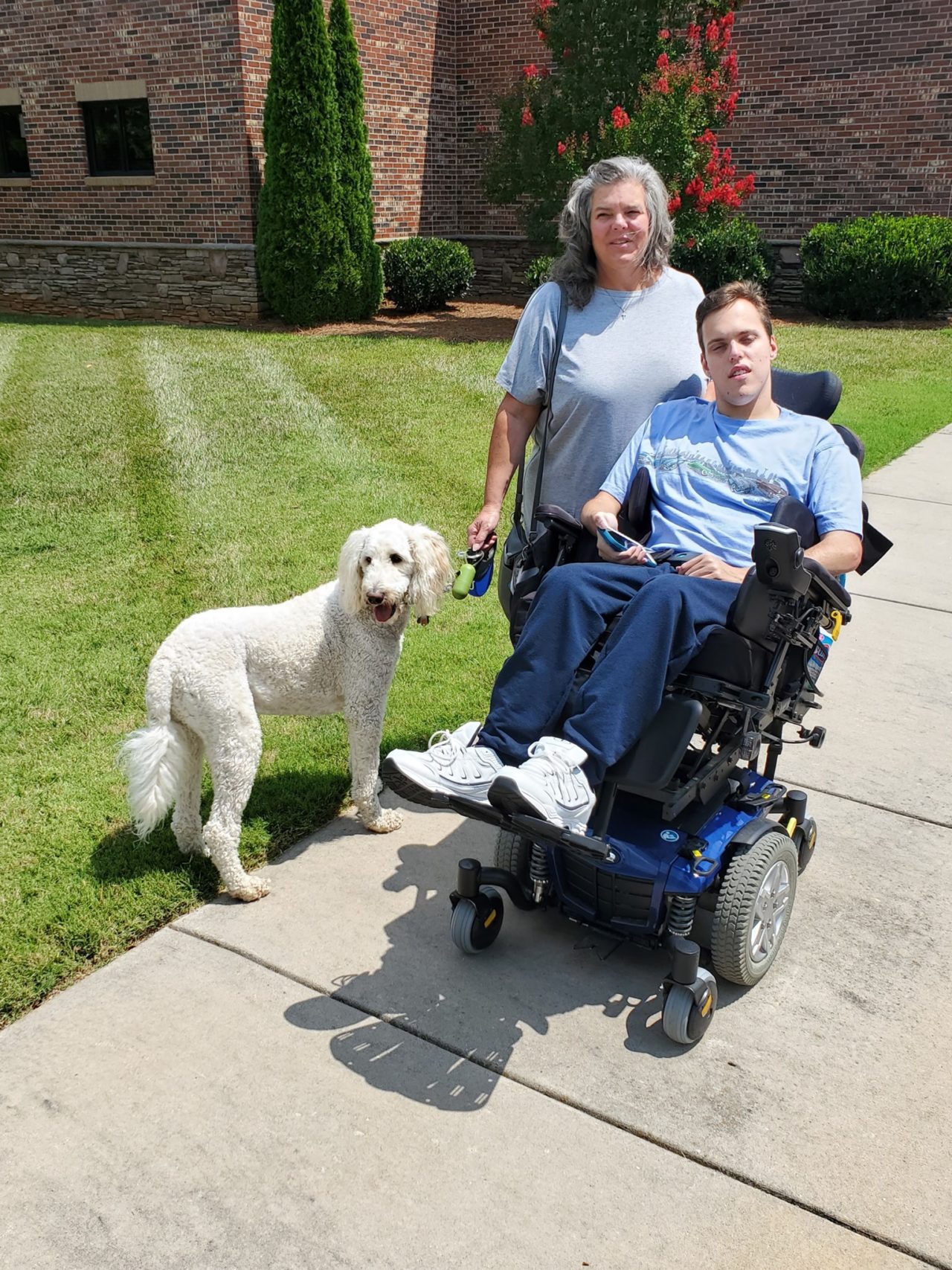 A mom stands beside a young man in a wheelchair. A white dog stands beside them.