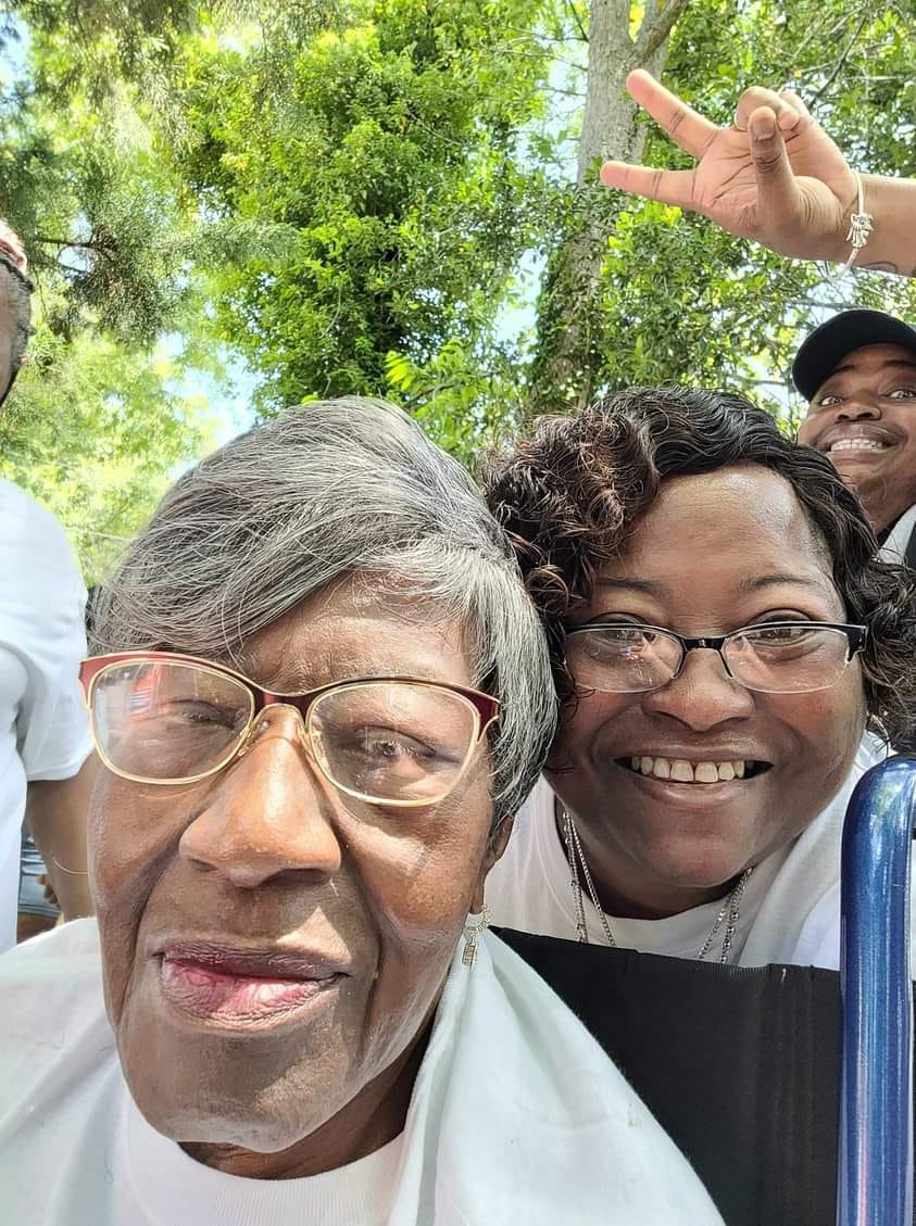 Two Black women smile, one older with gray hair and the other younger with short curly hair. A laughing young man photobombs from behind.
