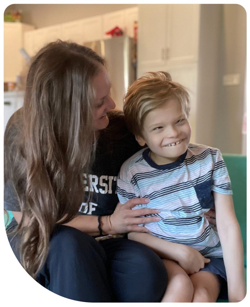Melissa and her son, Christopher. They are sitting next to each other with their bodies facing the camera. Melissa is looking at Christopher, her long hair partially obscuring her face, and her hands holding on to him. Christopher is smiling in the direction of the camera.