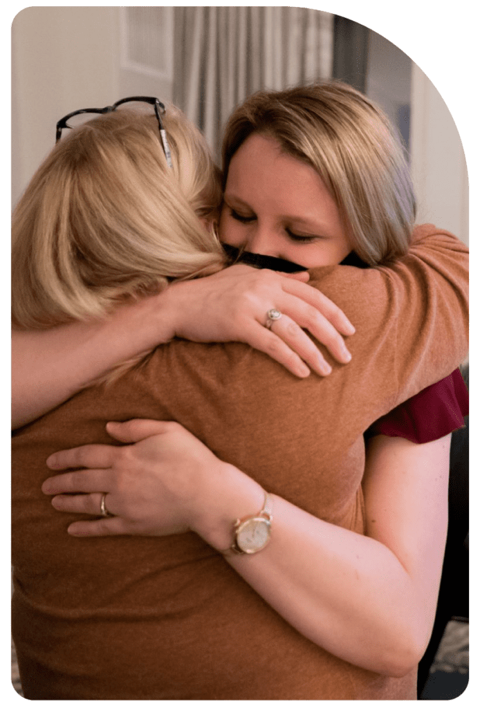 Holly Carmichael hugs another woman tightly. Both are wearing reddish tops and have blond hair.