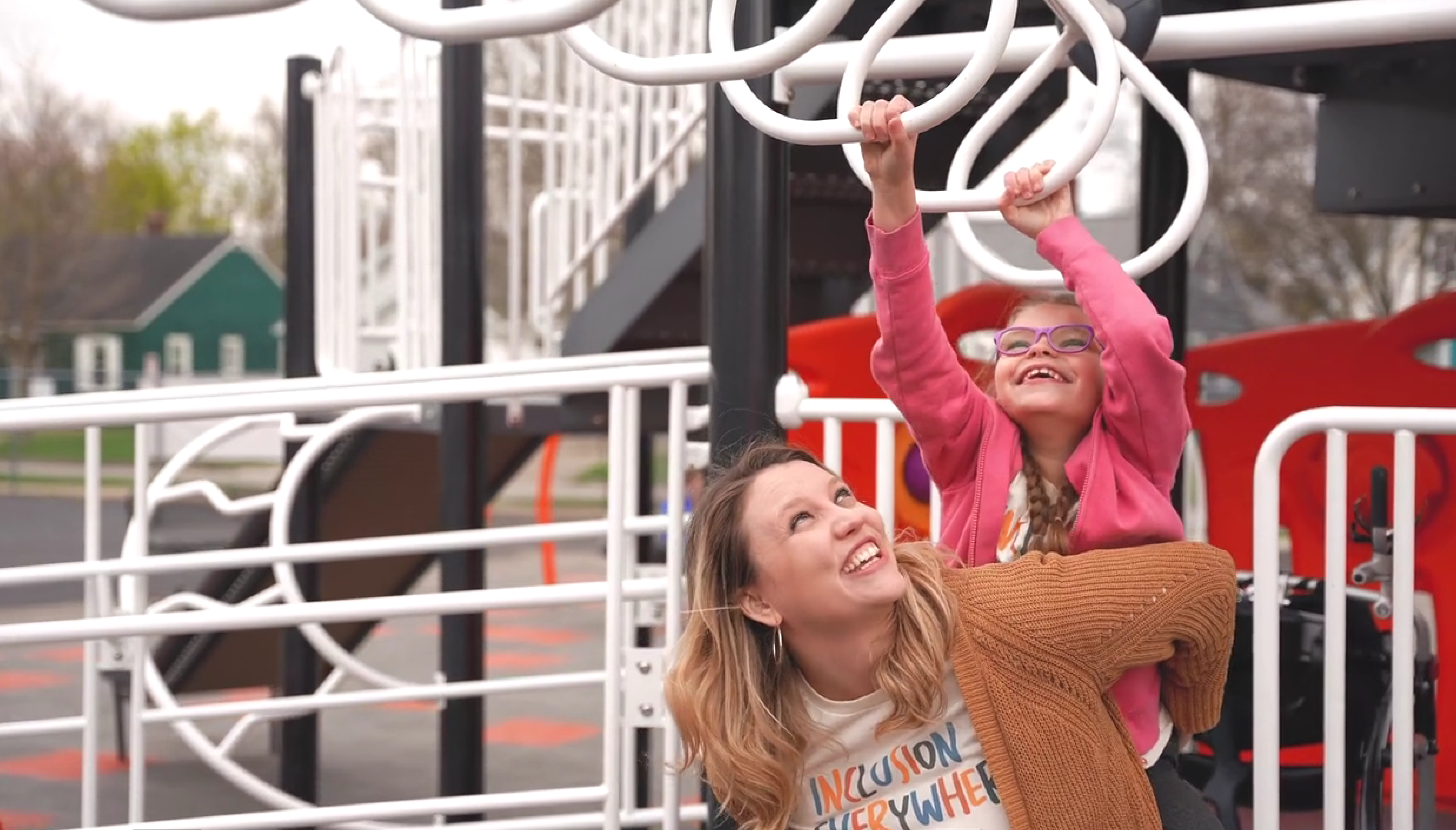 Holly holds Maggie on her back so she can reach the jungle gym bars. Both are wearing matching shirts that say "inclusion" and smiling.