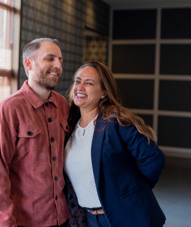 Nohora and Alex hug in the GT Office in front of a row of windows. Both are smiling. Alex wears a red button up and Nohora wears a white shirt with a navy blazer.