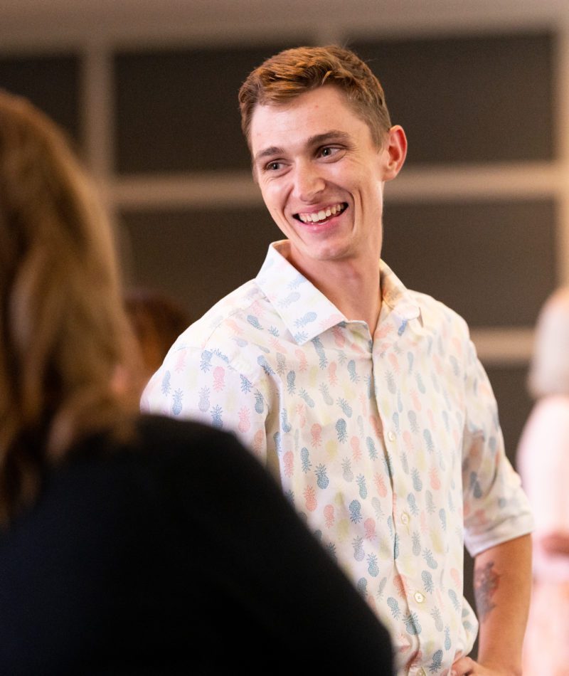 A customer service support specialist chats with coworkers at a company event. He is a white male with short brown hair and wears a shortsleeve button-up with a pineapple pattern. He smiles as he chats with a female coworker who's back is facing the camera.