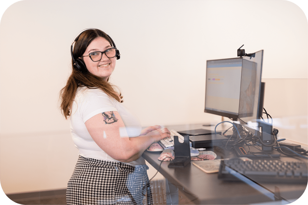 A woman with brown hair and glasses is smiling over her shoulder while standing in front of her computer at a standing desk. She is wearing a white shirt and houndstooth patterned pants.