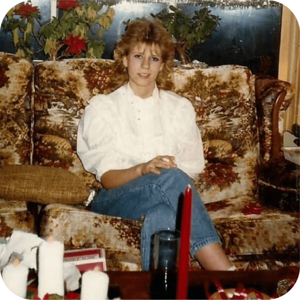 Kathy, seated on a large sofa with a 1970's pattern, is smiling at the camera. Her blonde hair is in a fashionable 1980's style, with bangs and large waves. She's wearing a white blouse and blue jeans.