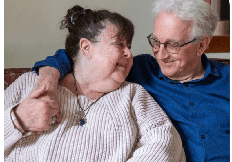 An older couple on the sofa smiling. The man has his arm wrapped around the woman, and she looks up at him with a big smile. The couch has colorful cushions.