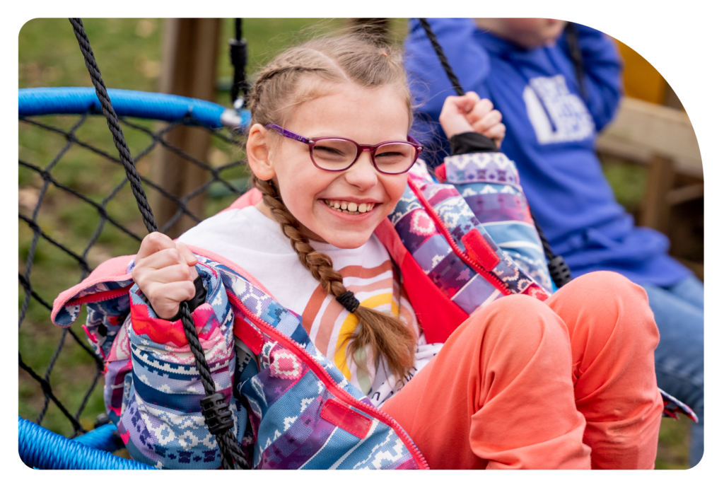 Maggie sits in an accessible swing at a playground, swinging herself with a huge smile. She wears a colorful jacket over a cream top and peach pants.