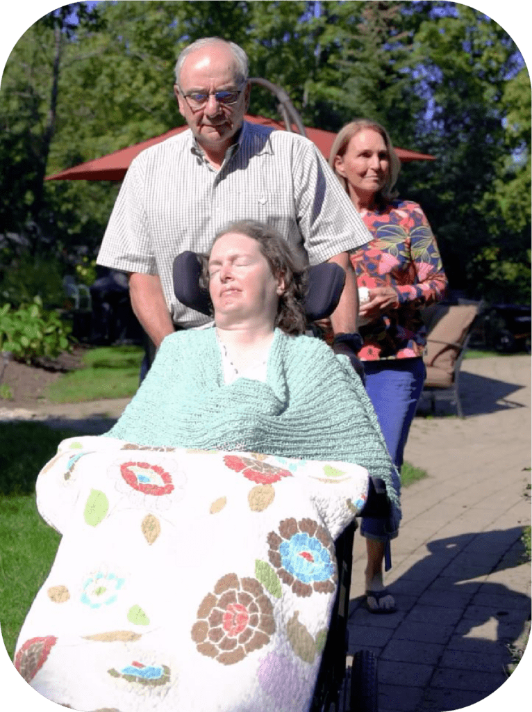 Jennifer being wheeled through the backyard by her father, Scott. Her mother, Mary, follows closely behind. The sun is shining, but it's still chilly, so Jennifer is under a quilted blanket.