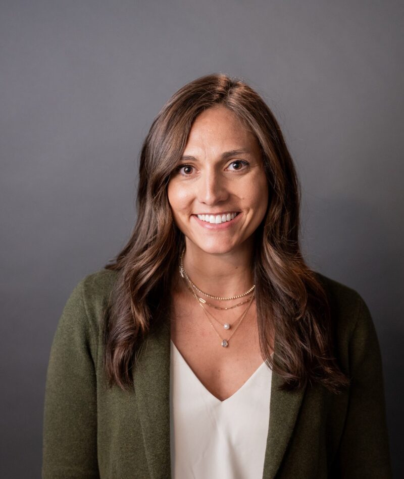 Kate smiles at the camera. She has wavy brown hair and a beautiful smile. She wears a green jacket over a white blouse.