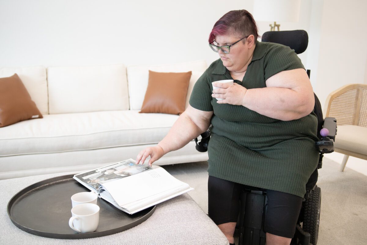 A woman in an automatic chair sits in the living room, looking through a helpful book. She has short brown hair and a long green tunic shirt. She's wearing black pants.