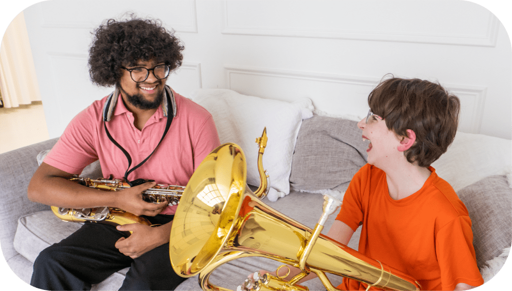 A young boy takes a band lesson from a younger man. They both sit on a gray couch. The boy holds a brass tuba on his lap and laughs at something his teacher says.