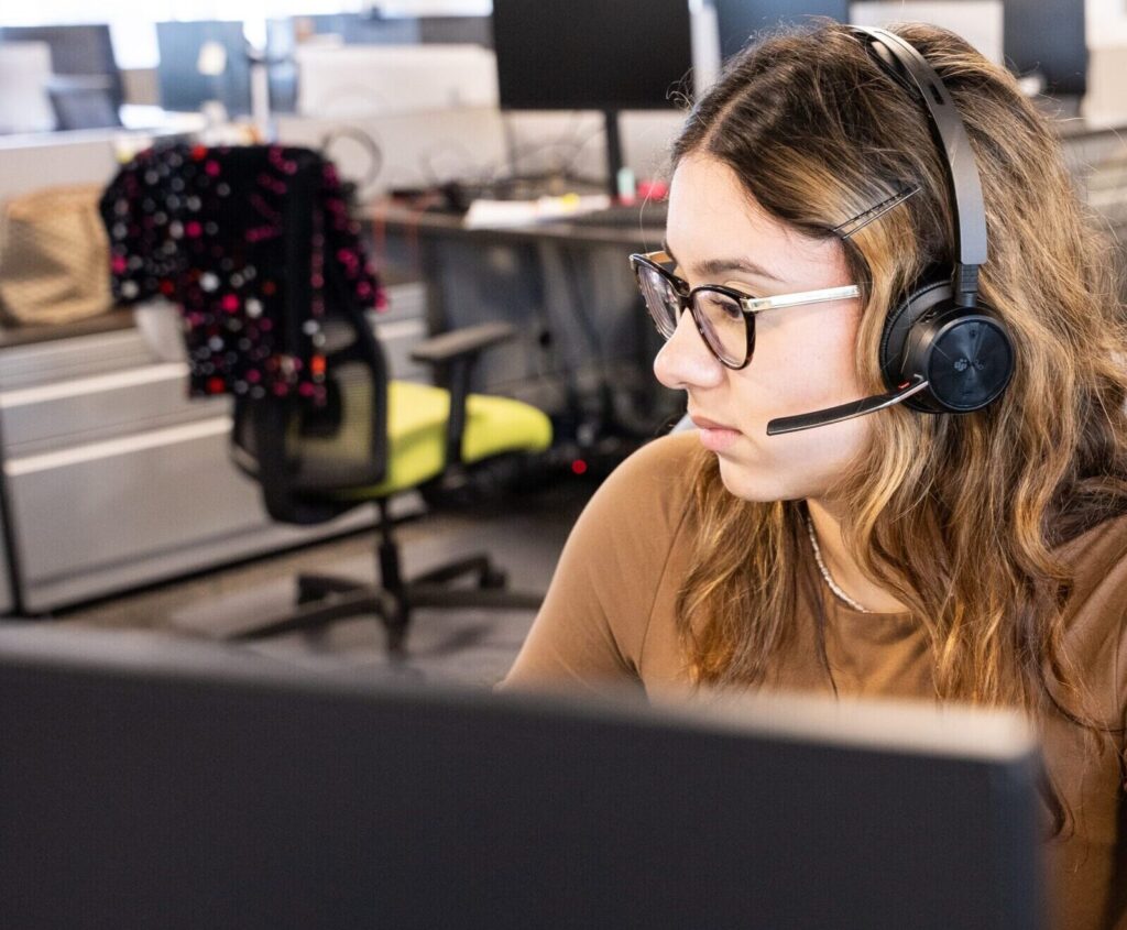 A woman works at a computer in the GTI office. She is a white woman with shoulder-lenth brown hair. She wears a light brown shirt, a black headset, and black-framed glasses.
