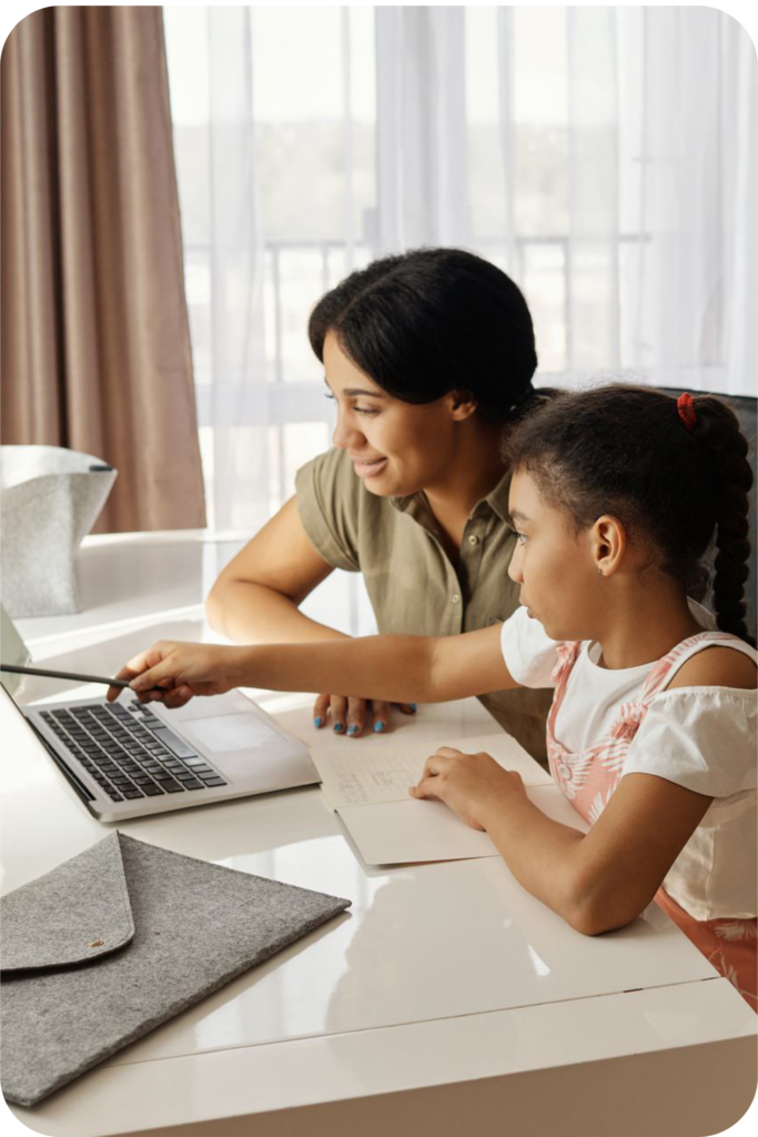 A mom and young daughter sit at the table in front of a laptop. The daughter, as the EOR, points at the laptop with a pen. Her other hand holds open a notebook with notes written inside. 