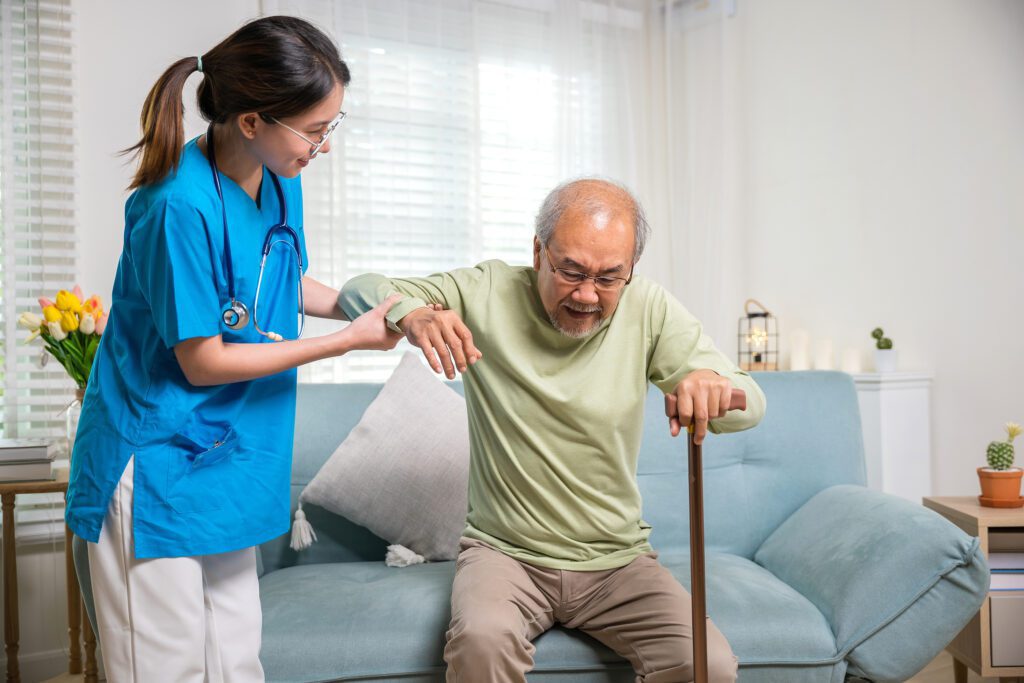 A man who uses a cane gets up from a couch with the assistance of a personal care attendant using self-direction services